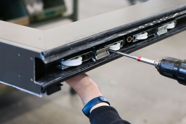 Een donker aluminium schuifraamkader met wieltjes wordt in de werkplaats vastgeschroefd